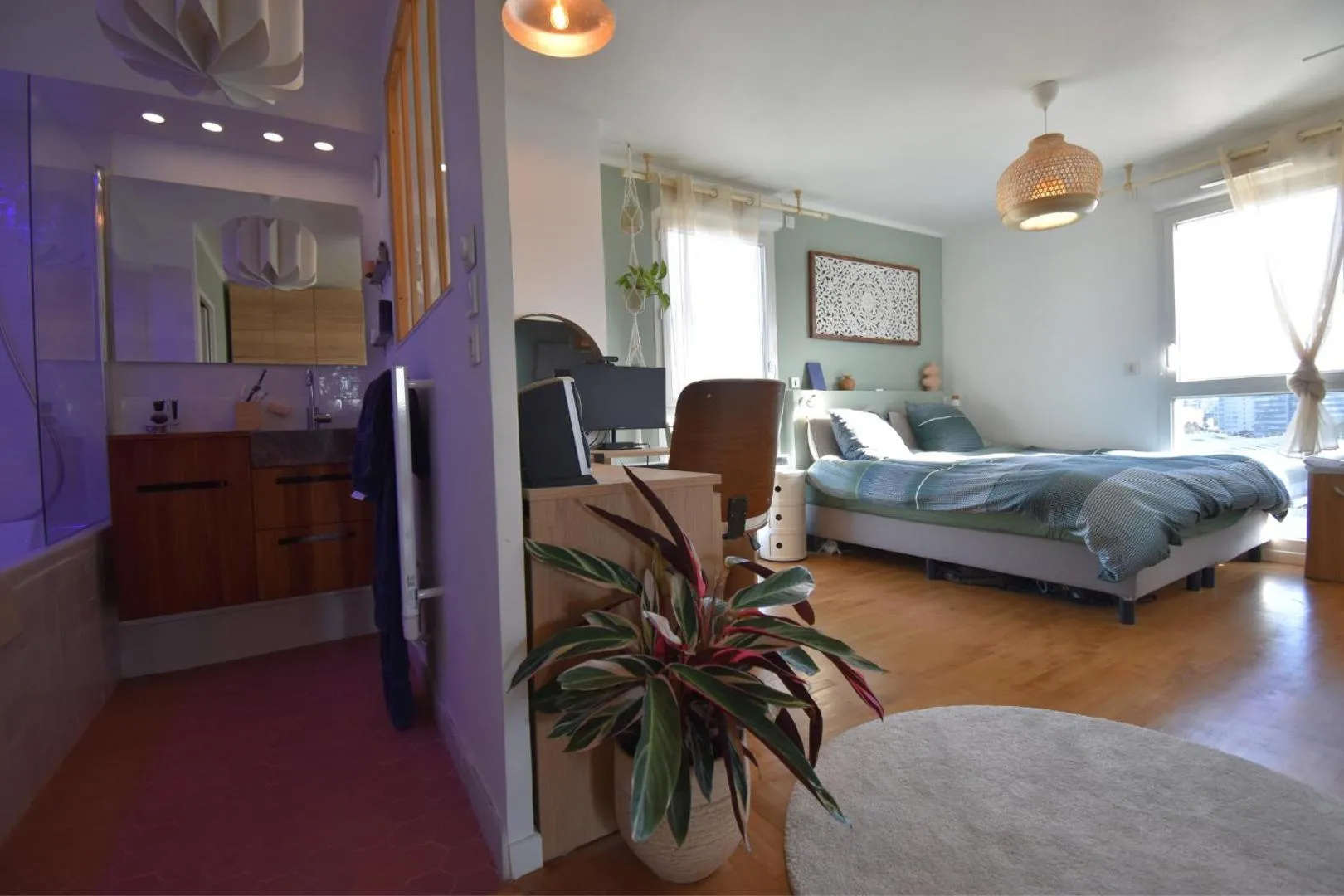 Chambre après : murs vert d'eau, lit gris clair, suspension en rotin, claustra en bois, salle de bain attenante à éclairage ambiant violet, plante tropicale au premier plan