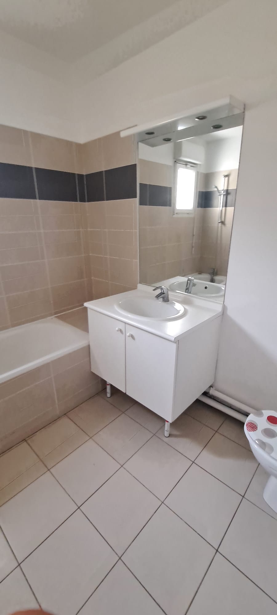 Salle de bain avant : carrelage beige avec liseré gris, baignoire standard, meuble blanc sur pieds, éclairage plafond
