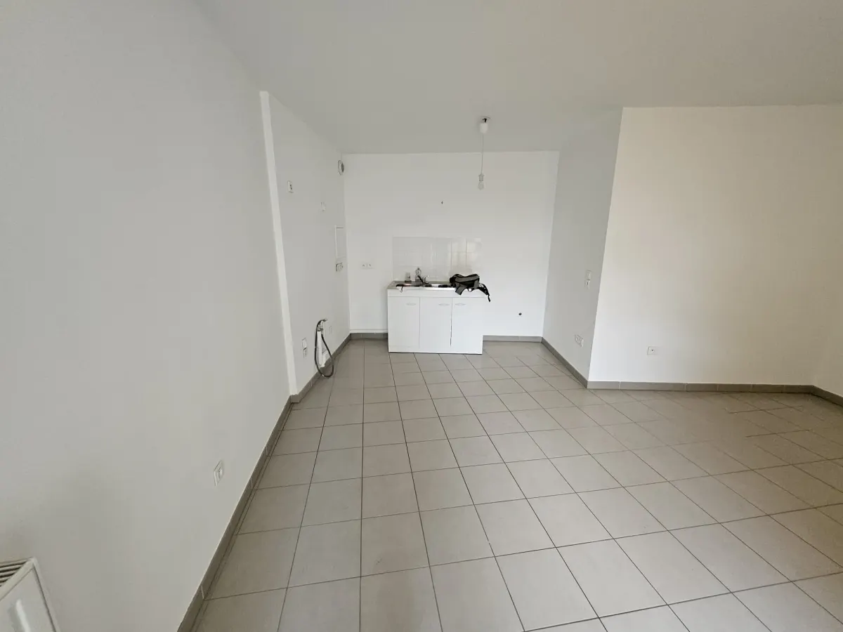 Séjour vide : carrelage gris, murs blancs, mini-kitchenette dans le fond, ampoule nue au plafond