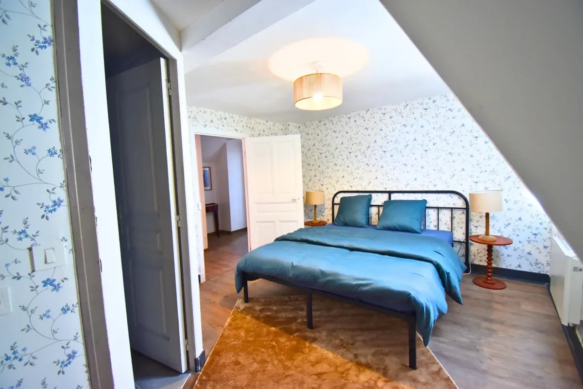 Chambre après home staging : papier peint fleuri bleu, lit en fer forgé noir, literie sarcelle, tables de nuit en bois tourné, tapis moutarde
